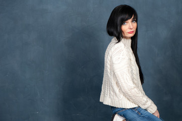 Fototapeta na wymiar Beautiful girl with long, black hair in a white knitted sweater and jeans is on her knees and sadly looks into the camera, a space for text. Studio portrait on a monochrome blue, trendy shabby back