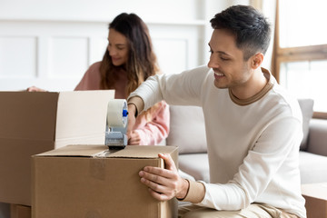 Smiling husband sealing box with adhesive tape, moving day concept, happy young couple packing...
