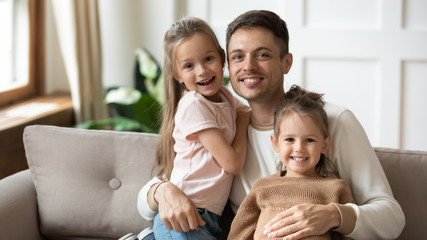 Fototapeta premium Head shot portrait happy father hugging two little daughters, sitting on couch at home, smiling young dad with preschool cute girls looking at camera, posing for family photo together