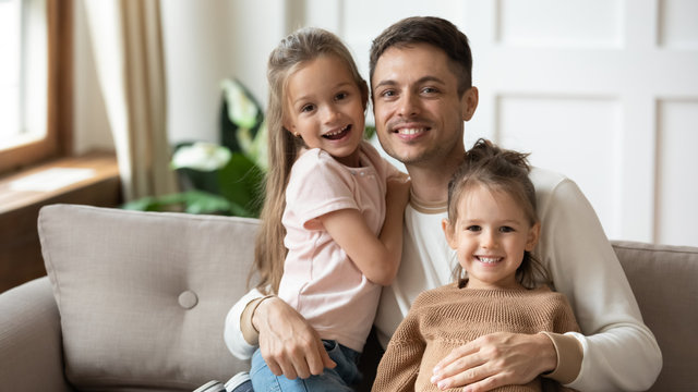Head shot portrait happy father hugging two little daughters, sitting on couch at home, smiling young dad with preschool cute girls looking at camera, posing for family photo together