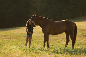 Teenaged girl stay with chestnut horse on pasture. Summer sunset on meadow, romantic scene