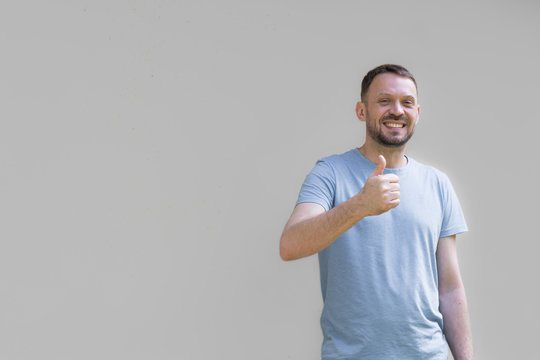 Uśmiechnięty mężczyzna na szarym tle, pokazujący dłonią gest ok, uniesiony w g&oacute;rę kciuk