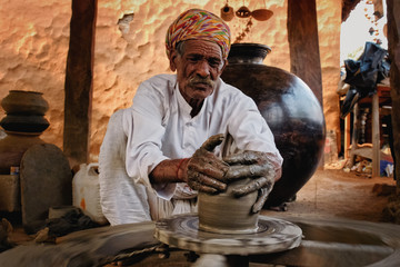 Indian potter at work: throwing the potter's wheel and shaping ceramic vessel and clay ware: pot, jar in pottery workshop. Experienced master. Handwork craft from Shilpagram, Udaipur, Rajasthan, India - obrazy, fototapety, plakaty