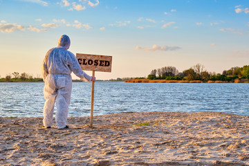 Man wearing protective suit stands with warning sign in hands with inscription closed along the sand beach of the river. Contaminated water, quarantine, virus outbreak, crime scene concept