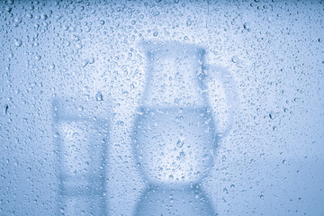 Pitcher with drinking water and a glass with a drink behind a glass with drops of water