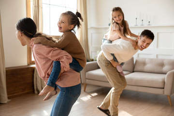 Happy parents with preschool children playing funny game, piggy back riding in living room, excited mother and father carrying two little daughters, running and laughing, enjoying leisure activity