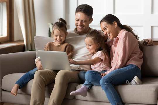 Happy family with two little daughters using laptop together, sitting on cozy sofa at home, smiling father and mother with preschool girls sisters looking at computer screen, shopping online - Powered by Adobe