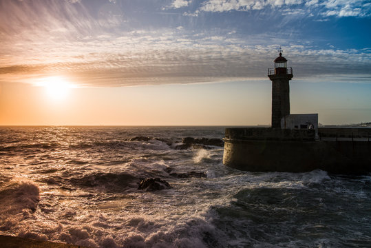 Paisaje marino: puesta de sol sobre un faro con la mar embravecida y nubes altas
