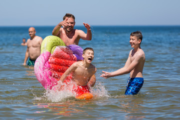 Zabawa w morzu, roześmiana rodzina chlapie się wodą, chłopiec spada z kolorowego materaca © Joanna