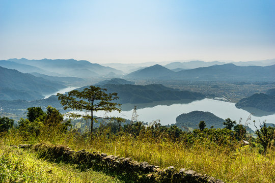 Panoramic view of Begnas and Rupa lakes from a hill above in Pokhara, Nepal, with layered hills, a stone wall, and a tree in the foreground under hazy atmospheric conditions