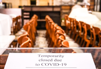 A restaurant closed announcement sign wording "temporarily closed due to COVID-19" with blurred table and chairs covered by white cloth background. Businesses are forced to shut down