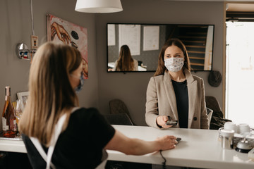 A girl who wears a medical face mask uses a cell phone to pay by contactless NFC technology. A female barista in a face mask holds out to a client a terminal for paying.