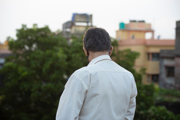 Back portrait of an Indian old man with corona preventive mask on a rooftop during sunset in home isolation.Indian lifestyle, disease and home quarantine.