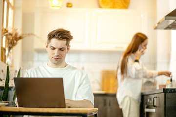Fototapeta na wymiar young caucasain married couple busy with their own business at home, man work on laptop from home while his wife make breakfast in the background. freelance, quarantine concept