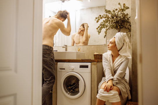 happy married caucasian couple spend morning in toilet together, woman sit on the pan in toilet after shower while man look at mirror, tidies up himself