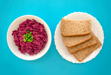 Tasty beet salad in a white bowl with brown bread on light blue background. Beetroot with cheese, sour cream and green onion, healthy and fresh food. Top view 