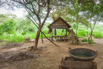 Myanmar Travel Images, Myanmar travel image of rustic stick and palm frond shelter in dirt patch under trees with washing stad with water bowl.