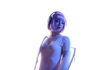 Beautiful woman with purple hair in futuristic costume over white background. Blue and violet neon light. Portrait of young girl in modern headphones listening music. Free space for text.