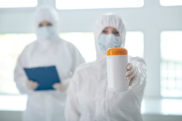 Medical worker in protective clothing showing disinfecting wipes bottle, her colleague standing behind with clip folder