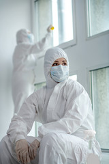 Medical worker in protective clothing and medical mask sitting next to the window, her collegue desinfecting panes