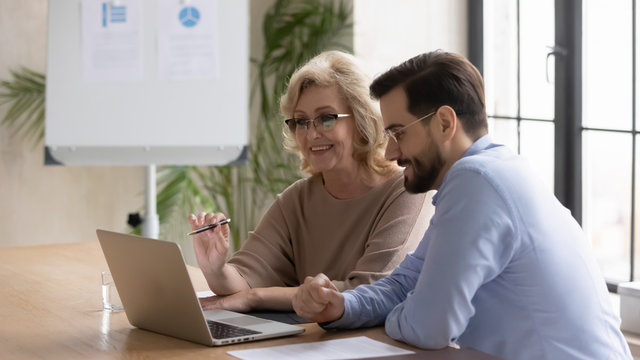 Smiling Mature 60 Year Old Businesswoman Using Laptop With Young Businessman Looking At Screen In Office At Meeting. Happy Woman Managers Talking About New Business Concept With Male Partner.