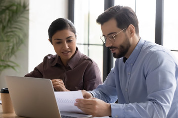 Diverse smiling businessman and businesswoman reading finance report talking looking at document. Indian woman and male manager discuss and looking decision in negotiation. Teamwork at workplace.