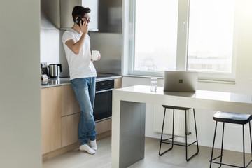 Young man talking on the phone and drinking coffee or tea while standing in the kitchen at home