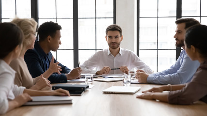 Serious businessman listening mature employee about project sit at table in boardroom at company meeting. Confident leader discuss business strategy with diverse colleagues in conversation.