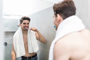 Health care, dental hygiene, people and beauty concept. Smiling young man with toothbrush cleaning teeth and looking to mirror at home bathroom