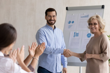 Obraz premium Happy 60 year old businesswoman congratulation hand shaking young businessman after success flipchart presentation new project in boardroom at meeting. Mature manager shaking hand good employee.