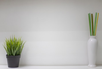 A black pot with green artificial grass and a white vase with bamboo colored sticks. They stand on a white shelf against a white wall. There is an empty space for writing text.