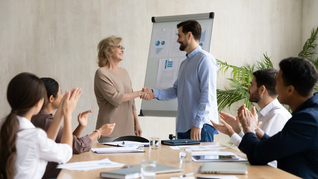 Happy young businessman congratulation hand shaking 60 year old businesswoman after success flipchart presentation new project in boardroom at meeting. Manager shaking hand mature good employee. - Powered by Adobe