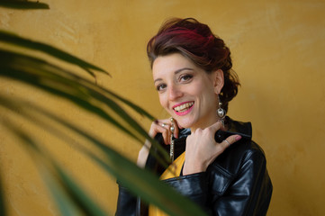 Glamorous lady in punk rock style party clothes with dark pink hair in casual dress, black leather jacket, and long earrings on yellow background