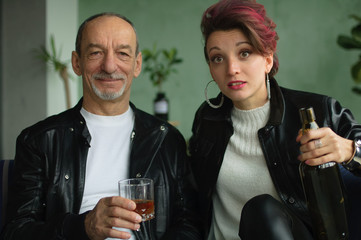 Indoor studio shot of excited beautiful woman with dark pink hair in white sweater and black leather jacket drinking with her senior father in loft style room. Female alcoholism