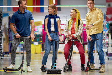 positive caucasian sportive men and women use scooters in aisle of store, try it, going to make purchase, customers have fun, laugh while shopping together