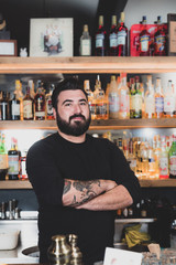 Young bartender smiles at you while you're at the counter, background shelves with bottles of spirits and liquors