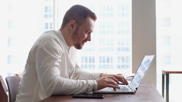Side view of cheerful bearded young businessman wearing fashion casual clothing working on laptop computer at the desk in modern office room on background of large window, mobile phone on table.
