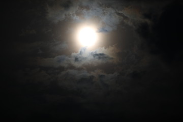 Beautiful night sky background. An indistinct image of a full moon in the dark in a fog surrounded by gray, pale-lit fluffy clouds of gloomy tones.The mystical picture
