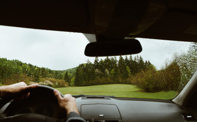 Driver driving a modern off road left hand drive LHD car on the mountain green forest country road at rainy moody day. POV inside car windshield view point. Safely auto driving concept.