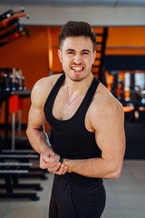 Fototapeta premium Young muscular man screaming and showing his bicep with angry face in the gym. Wearing black shirt and black sweatpants. Screaming man with well trained body, biceps, abs and pecs.