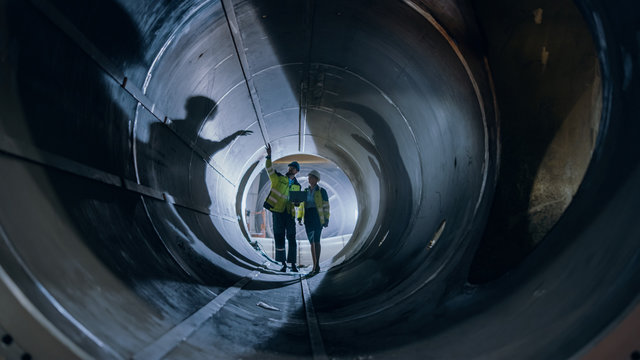 Two Heavy Industry Engineers Walking Inside Pipe, Use Laptop, Have Discussion, Checking Welding. Construction of the Oil, Natural Gas and fuels Transport Pipeline. Industrial Manufacturing Factory