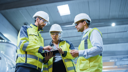Three Heavy Industry Engineers Stand in Pipe Manufacturing Factory, Use Digital Tablet Computer,...