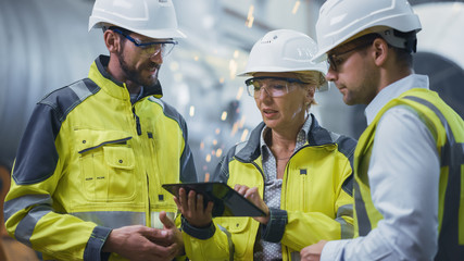 Three Heavy Industry Engineers Stand in Pipe Manufacturing Factory, Use Digital Tablet Computer,...