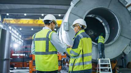 Two Heavy Industry Engineers Walk Through Pipe Manufacturing Factory, Use Digital Tablet Computer,...