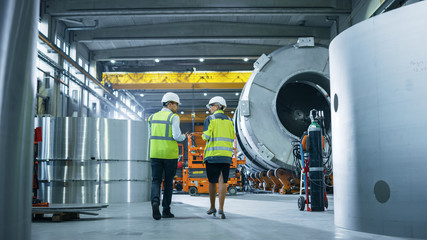 Two Heavy Industry Engineers Walk Through Pipe Manufacturing Factory, Use Digital Tablet Computer, Have Discussion. Facility for Construction of Oil, Gas and Fuel Pipeline Transportation Products