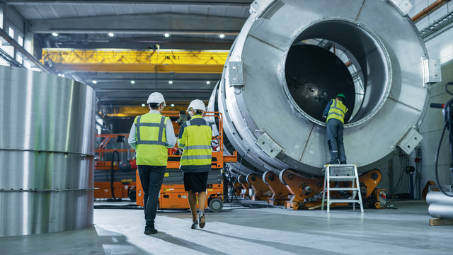 Two Heavy Industry Engineers Walk Through Pipe Manufacturing Factory, Use Digital Tablet Computer, Have Discussion. Facility for Construction of Oil, Gas and Fuel Pipeline Transportation Products