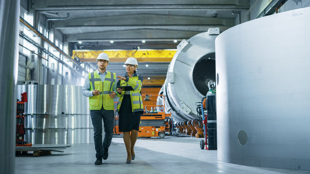 Two Heavy Industry Engineers Walk Through Pipe Manufacturing Factory, Use Digital Tablet Computer, Have Discussion. Facility for Construction of Oil, Gas and Fuel Pipeline Transportation Products.