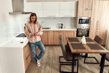 Getting ready. Young woman using red marijuana grinder, standing in the kitchen. Glass water pipe or bong, cannabis buds in a plastic bag on the table. Cannabis and weed legalization concept