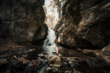 Woman (traveler and adventure) standing under the waterfall in dark and moody forest - surounded by stones and rocks. Folk tone shot of woman looking on the waterfall with backpack and relaxing.