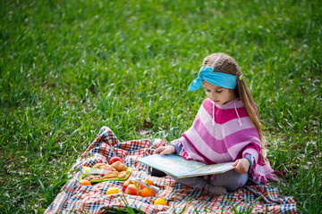 little girl child sits on a bedspread and reads a book with a fairy tale, green grass in the field, sunny spring weather, smile and joy of the child, blue sky with clouds